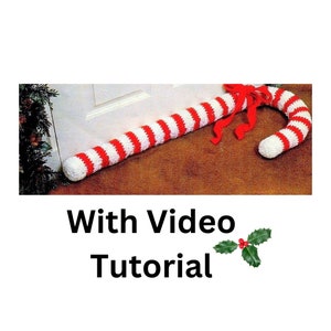 Puede incluir: Un tapón de puerta de caramelo tejido a crochet con rayas rojas y blancas con un lazo de cinta roja. El tapón de puerta está colocado en la parte inferior de una puerta blanca. El texto "With Video Tutorial" y una rama de acebo están debajo de la imagen.