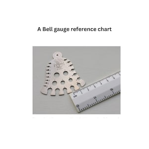 May include: A silver bell-shaped gauge with numbered holes for measuring different sizes. The gauge is next to a ruler marked in centimeters.