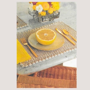 May include: A beige crocheted placemat featuring a round ceramic plate with a halved yellow grapefruit, a gold fork, a gold spoon, and a clear glass vase filled with yellow and white flowers, all arranged on a marble surface.