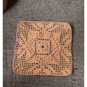 May include: A square crocheted doily in a light brown color with a detailed pattern. The doily has a center square with a smaller circle in the center. The doily is made with a single crochet stitch.