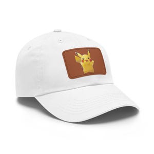 Op de afbeelding: Een witte baseballpet met een rechthoekige bruine patch op het voorpaneel. De patch toont een afbeelding van het gele personage Pikachu met rode wangen, puntige oren met zwarte uiteinden en een kleine glimlach, staand met opgeheven armen.
