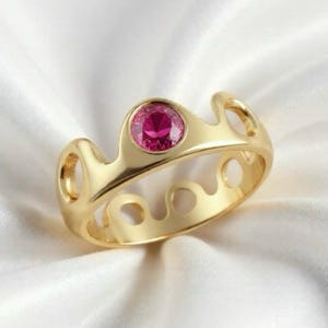 May include: A gold-colored ring with a round, bright pink gemstone set in the center. The ring has a unique openwork design with curved shapes and circular cutouts. The ring is displayed on a white, silky fabric.