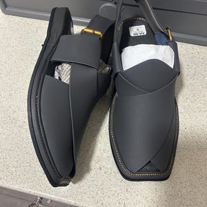 May include: A pair of gray leather sandals with a strap and buckle closure. The sandals have a stitched sole and a square toe design. The sandals are size 9.