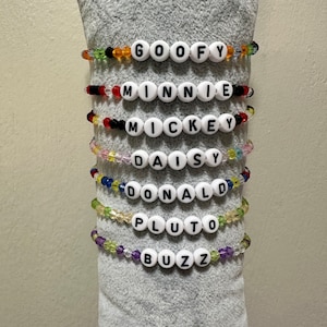 May include: Seven colorful beaded bracelets with white letter beads spelling out the names "GOOFY", "MINNIE", "MICKEY", "DAISY", "DONALD", "PLUTO", and "BUZZ".