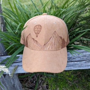 K&ouml;nnte beinhalten: Eine Baseballkappe aus braunem Wildleder mit einem braunen gestickten Design von Bergen und einem Hei&szlig;luftballon.
