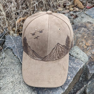 May include: A brown suede baseball cap with an embroidered design of mountains and birds. The cap has a curved brim and a mesh back.
