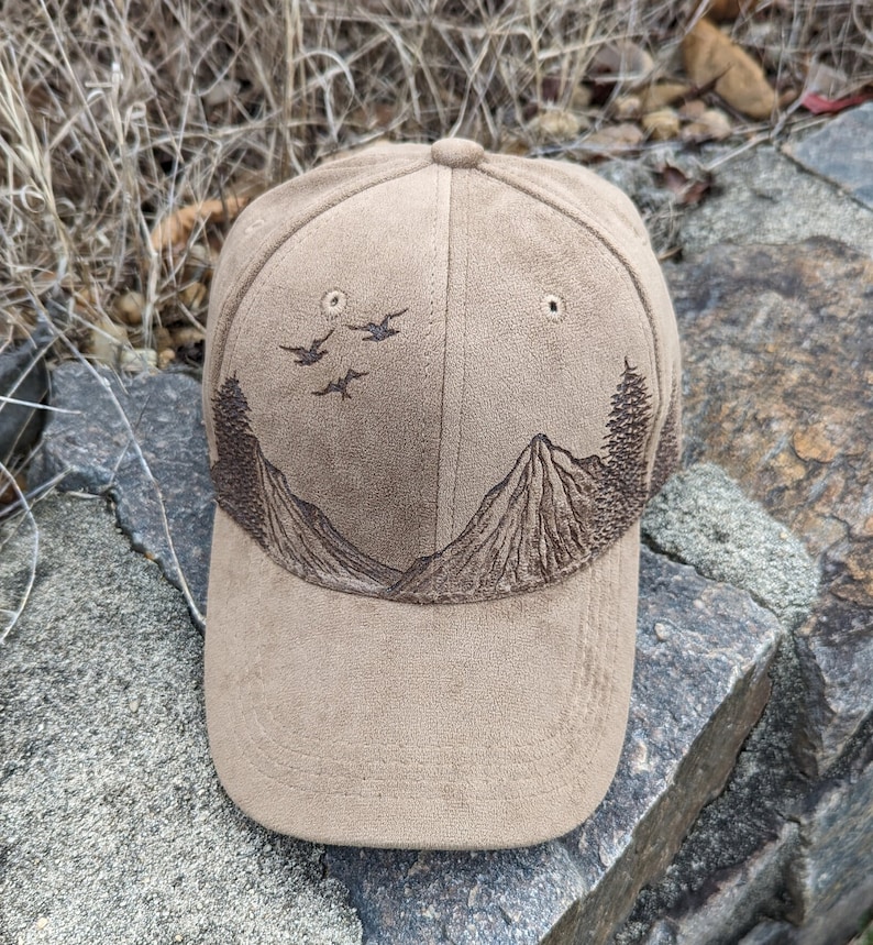 May include: A brown suede baseball cap with an embroidered design of mountains and birds. The cap has a curved brim and a mesh back.