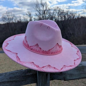 Könnte beinhalten: Ein rosafarbener Filzhut im Fedora-Stil mit einem rosafarbenen Bergmotiv und einem rosafarbenen Band um die Krone.