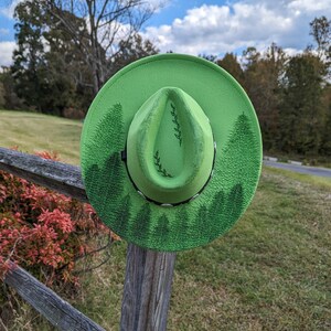 Puede incluir: Un sombrero fedora verde brillante con un dise&ntilde;o de bosque. El sombrero presenta una banda negra y una ilustraci&oacute;n detallada de pinos alrededor del borde. La corona del sombrero tiene un dise&ntilde;o de hojas simple.
