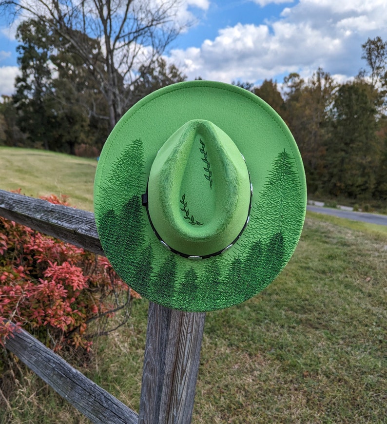Puede incluir: Un sombrero fedora verde brillante con un dise&ntilde;o de bosque. El sombrero presenta una banda negra y una ilustraci&oacute;n detallada de pinos alrededor del borde. La corona del sombrero tiene un dise&ntilde;o de hojas simple.