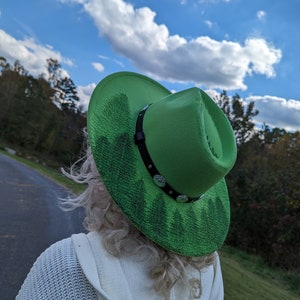 Puede incluir: Un sombrero de vaquero verde brillante con una banda de cuero negro y adornos plateados. El ala presenta un dise&ntilde;o de bosque pintado. El sombrero se usa al aire libre contra un cielo azul con nubes.