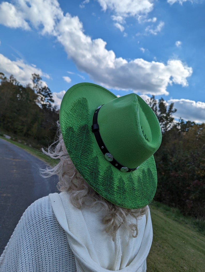 Puede incluir: Un sombrero de vaquero verde brillante con una banda de cuero negro y adornos plateados. El ala presenta un dise&ntilde;o de bosque pintado. El sombrero se usa al aire libre contra un cielo azul con nubes.