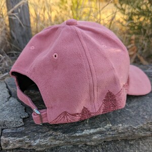 May include: A dusty rose colored baseball cap with an embroidered forest scene on the back. The cap has a black brim and a silver buckle.