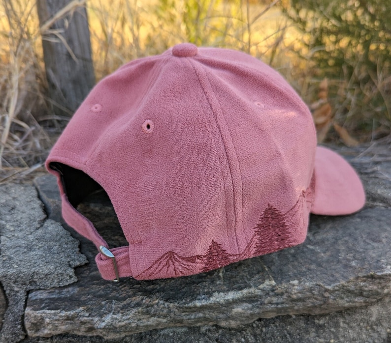 May include: A dusty rose colored baseball cap with an embroidered forest scene on the back. The cap has a black brim and a silver buckle.