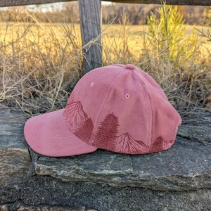 May include: A pink baseball cap with a forest scene embroidered on the front. The cap has a curved brim and a structured crown.