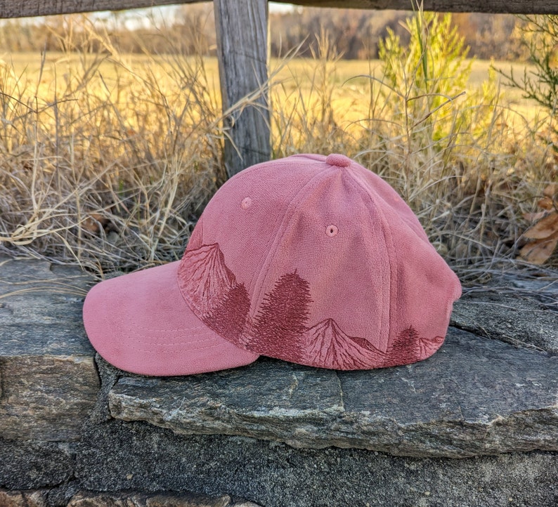 May include: A pink baseball cap with a forest scene embroidered on the front. The cap has a curved brim and a structured crown.