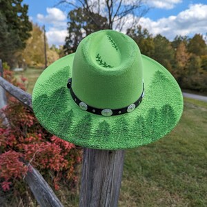 Puede incluir: Un sombrero fedora verde brillante con una banda negra y adornos plateados. El ala presenta un dise&ntilde;o de bosque, y la corona tiene un patr&oacute;n de hojas. El sombrero se exhibe al aire libre sobre un poste de madera.
