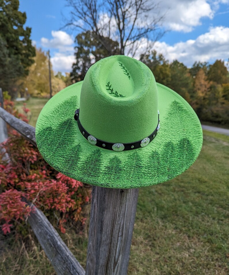Puede incluir: Un sombrero fedora verde brillante con una banda negra y adornos plateados. El ala presenta un dise&ntilde;o de bosque, y la corona tiene un patr&oacute;n de hojas. El sombrero se exhibe al aire libre sobre un poste de madera.