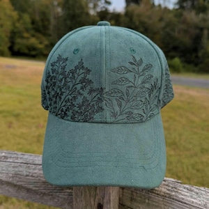 May include: A teal baseball cap with a curved brim. The cap features a black floral design on the crown. The hat is made of a soft material and is suitable for outdoor activities.