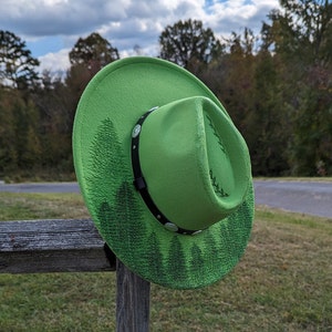 Puede incluir: Un sombrero de vaquero verde brillante con una banda negra y detalles plateados. El sombrero presenta un dise&ntilde;o de bosque en el ala y la corona. Descansa sobre un poste de valla de madera, con &aacute;rboles y un cielo nublado.