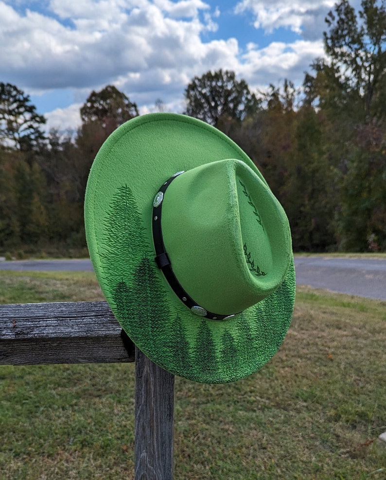 Puede incluir: Un sombrero de vaquero verde brillante con una banda negra y detalles plateados. El sombrero presenta un dise&ntilde;o de bosque en el ala y la corona. Descansa sobre un poste de valla de madera, con &aacute;rboles y un cielo nublado.