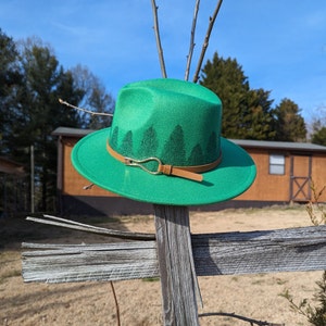 May include: A green felt fedora hat with a brown leather band and a gold buckle. The hat has a design of pine trees around the band.