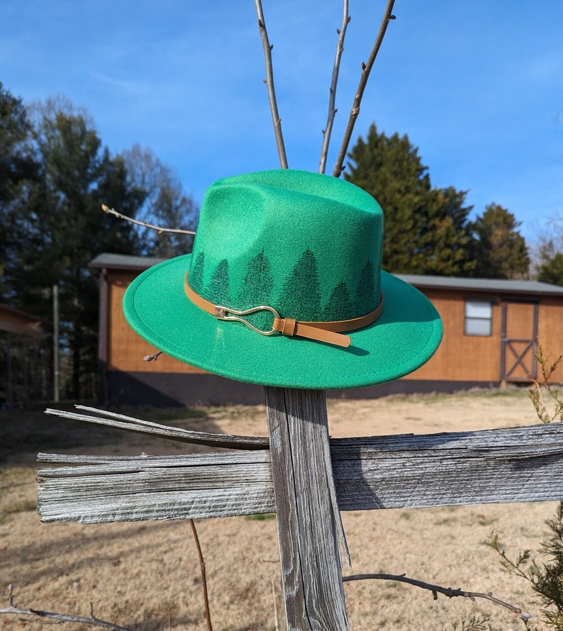 May include: A green felt fedora hat with a brown leather band and a gold buckle. The hat has a design of pine trees around the band.
