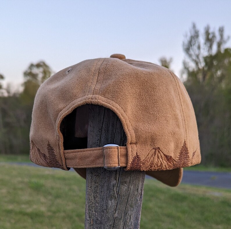 K&ouml;nnte beinhalten: Eine braune Baseballkappe aus Wildleder mit einem gestickten Bergpanorama auf der R&uuml;ckseite. Die Kappe hat einen verstellbaren Riemen in Braun mit einer silbernen Schnalle.