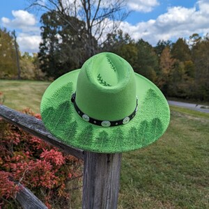 Puede incluir: Un sombrero de vaquero verde brillante con una banda negra adornada con tachuelas plateadas. El sombrero presenta un dise&ntilde;o de bosque alrededor del ala y hojas decorativas en la corona. El sombrero se muestra al aire libre en un poste de valla de madera.