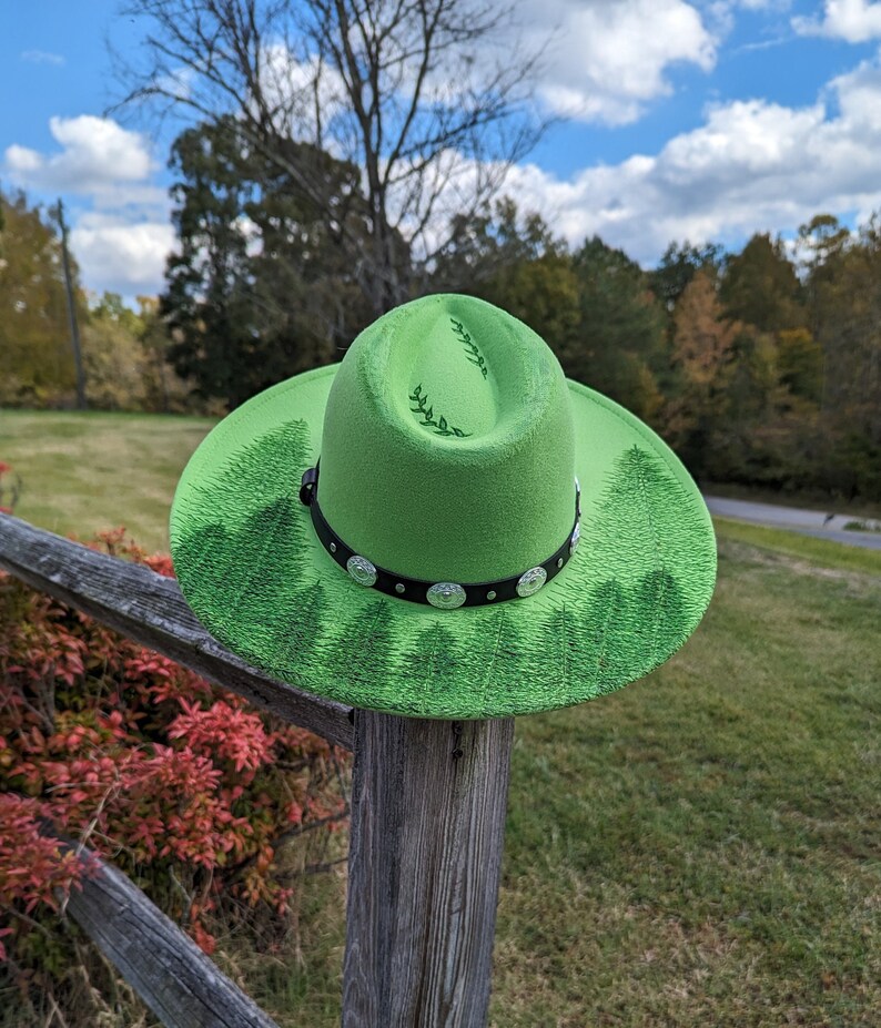 Puede incluir: Un sombrero de vaquero verde brillante con una banda negra adornada con tachuelas plateadas. El sombrero presenta un dise&ntilde;o de bosque alrededor del ala y hojas decorativas en la corona. El sombrero se muestra al aire libre en un poste de valla de madera.