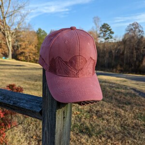 May include: A pink baseball cap with an embroidered design of mountains and a sun. The cap is sitting on a wooden fence post.