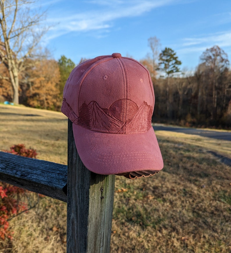 May include: A pink baseball cap with an embroidered design of mountains and a sun. The cap is sitting on a wooden fence post.