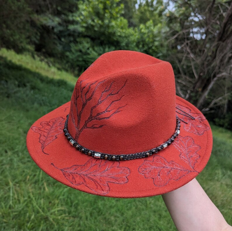 May include: A red felt fedora hat with a brown leather band and silver accents. The hat is decorated with a black and white design of oak leaves and branches.