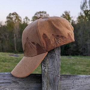 K&ouml;nnte beinhalten: Eine braune Baseballkappe aus Wildleder mit einem gestickten Bergpanorama in braunem Garn. Die Kappe sitzt auf einem Holzpfosten.