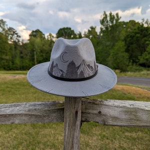 Op de afbeelding: Een grijze wollen fedora hoed met een zwarte band en een berg- en maanontwerp. De hoed ligt op een houten hek.