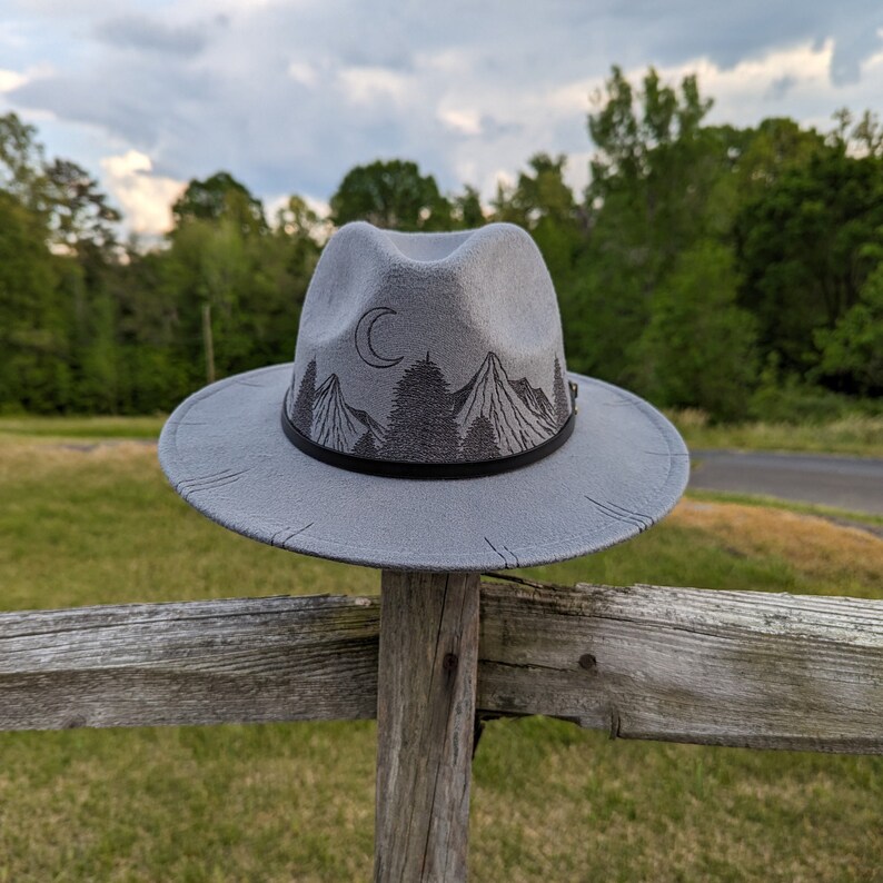 Op de afbeelding: Een grijze wollen fedora hoed met een zwarte band en een berg- en maanontwerp. De hoed ligt op een houten hek.