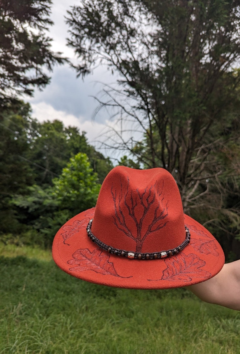May include: A red felt fedora hat with a black tree design and brown leather trim. The hat is decorated with brown beads and has a wide brim.