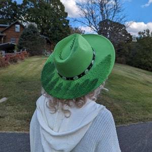 Puede incluir: Un sombrero de vaquero verde brillante con un dise&ntilde;o de bosque pintado en el ala. El sombrero tiene una banda negra con tachuelas plateadas. Una persona con un su&eacute;ter blanco lleva el sombrero.