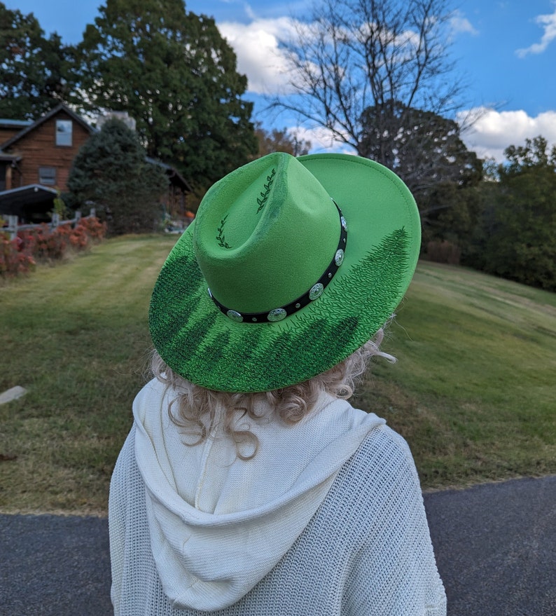 Puede incluir: Un sombrero de vaquero verde brillante con un dise&ntilde;o de bosque pintado en el ala. El sombrero tiene una banda negra con tachuelas plateadas. Una persona con un su&eacute;ter blanco lleva el sombrero.
