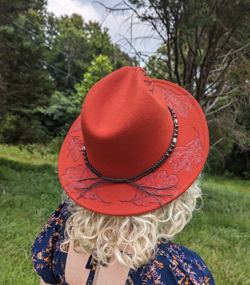 May include: A red felt fedora hat with a brown leather band and embroidered oak leaves. The hat has a wide brim and a slightly pointed crown.