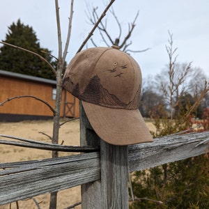 May include: A brown baseball cap with a mountain and birds design. The cap is sitting on a wooden fence post.