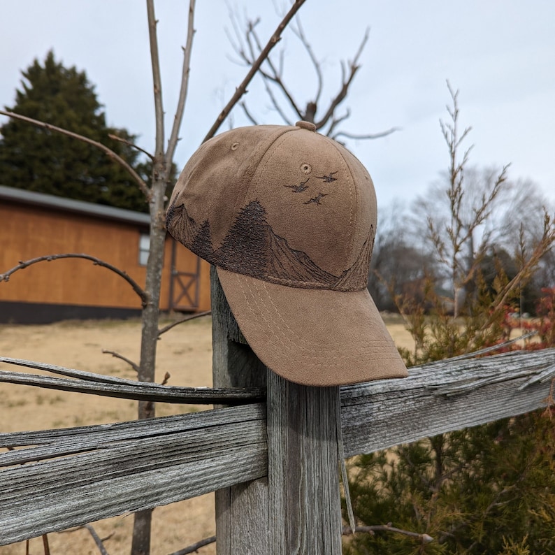 May include: A brown baseball cap with a mountain and birds design. The cap is sitting on a wooden fence post.