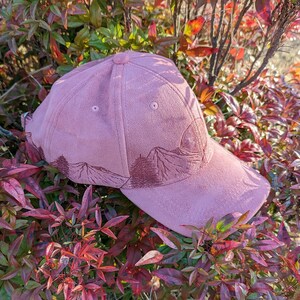 May include: A pink suede baseball cap with an embroidered mountain scene. The cap has a curved brim and a slightly distressed look.