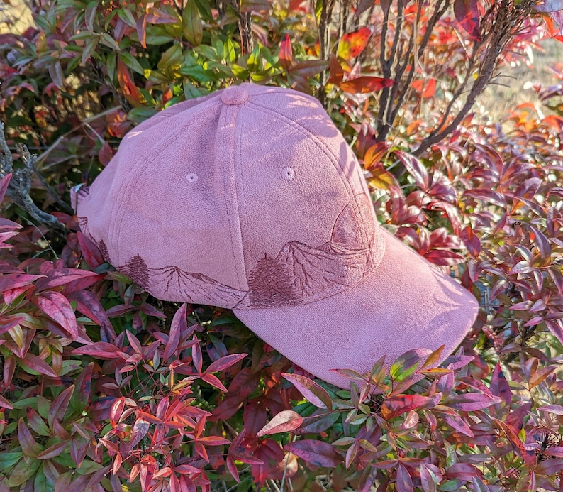 May include: A pink suede baseball cap with an embroidered mountain scene. The cap has a curved brim and a slightly distressed look.