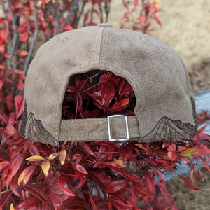 May include: A brown suede baseball cap with a mountain design embroidered on the back. The cap has a silver adjustable strap.