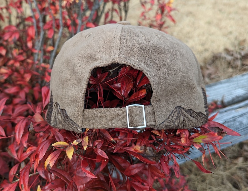 May include: A brown suede baseball cap with a mountain design embroidered on the back. The cap has a silver adjustable strap.