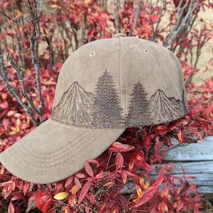 May include: A brown baseball cap with an embroidered design of mountains and trees. The cap has a curved brim and a brown adjustable strap.