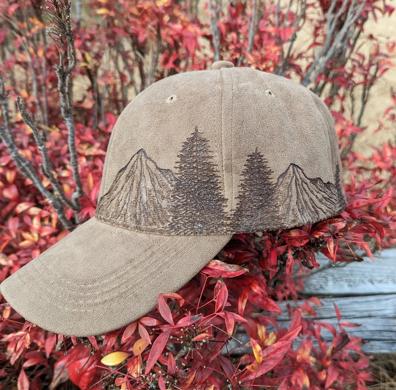 May include: A brown baseball cap with an embroidered design of mountains and trees. The cap has a curved brim and a brown adjustable strap.