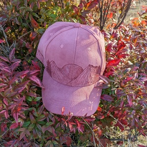 May include: A pink baseball cap with a mountain scene embroidered on the front. The mountains are brown and the sky is a light pink.
