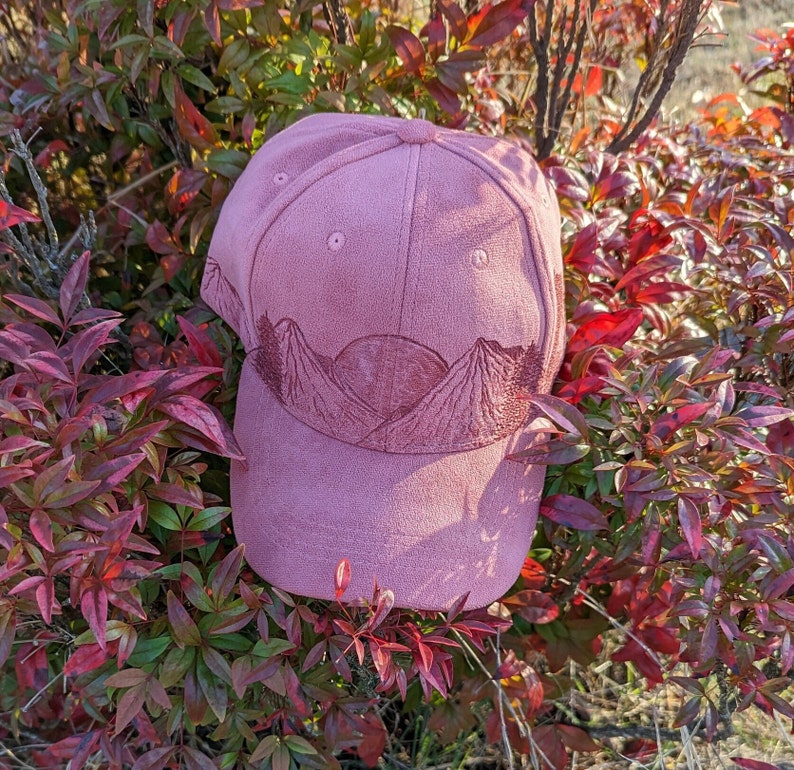 May include: A pink baseball cap with a mountain scene embroidered on the front. The mountains are brown and the sky is a light pink.